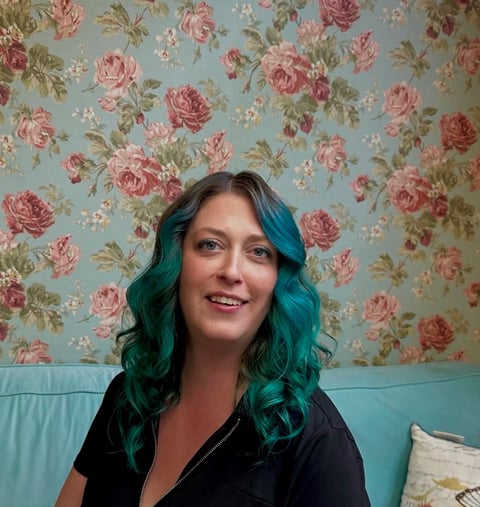 Woman with teal wavy hair and red lipstick smiling at camera, wearing black top against floral wallpaper and turquoise couch