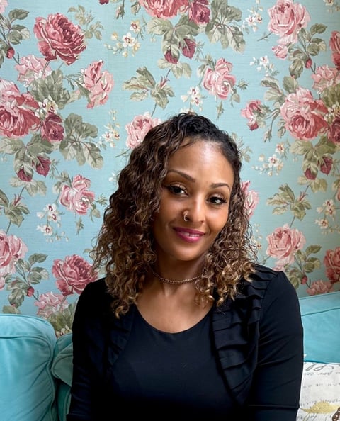 Woman with curly dark hair wearing a black top and silver necklace, smiling in front of a blue floral wallpaper background